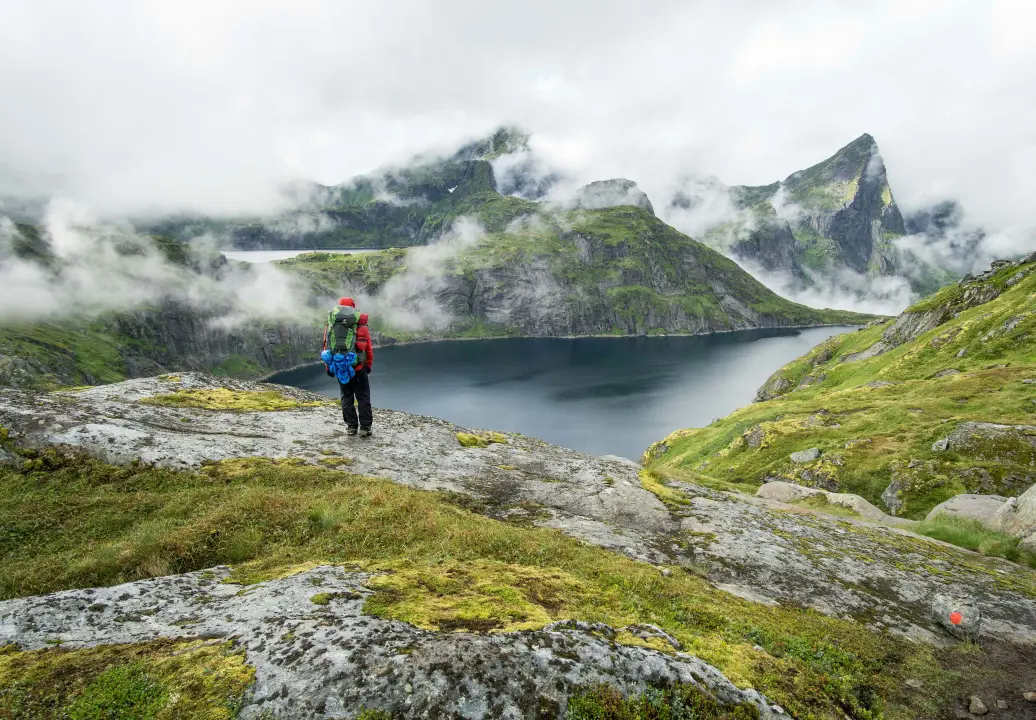 A hiker in a red jacket stands on rocky terrain overlooking a serene lake surrounded by lush green mountains and misty clouds.