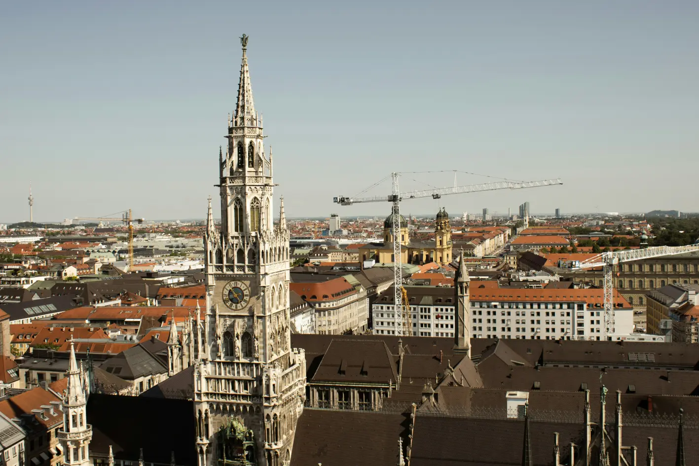 A panoramic view of Munich featuring the ornate tower of the New Town Hall, surrounding rooftops, and construction cranes in the background.