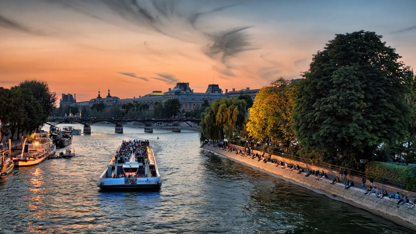 A scenic view of the Seine River at sunset, featuring a crowded boat, lush trees, and people sitting along the riverbank.