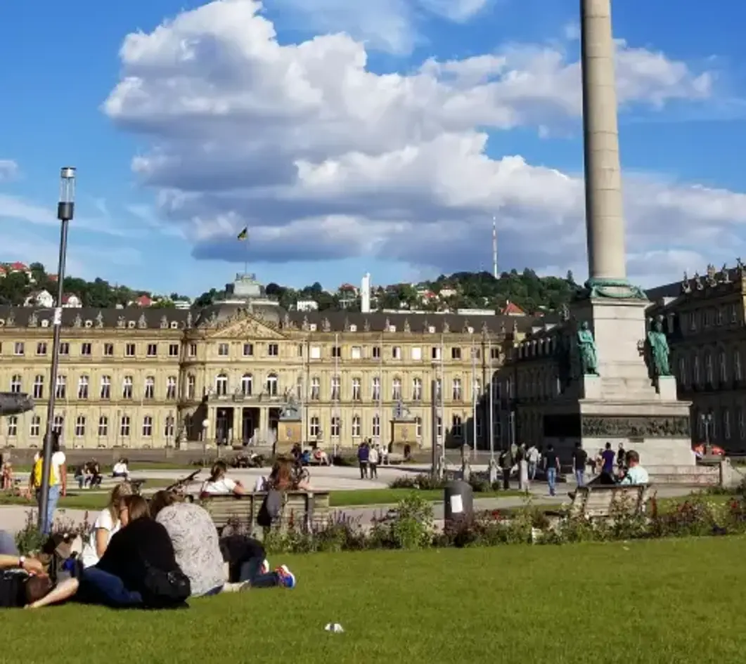 People relax on grass in front of a grand building with columns, under a blue sky with fluffy clouds and a tall monument nearby.