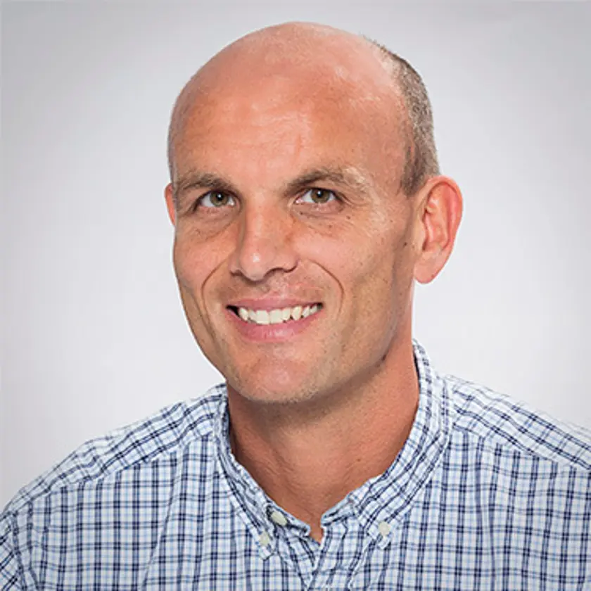 A smiling bald man wearing a checkered shirt poses against a neutral background.