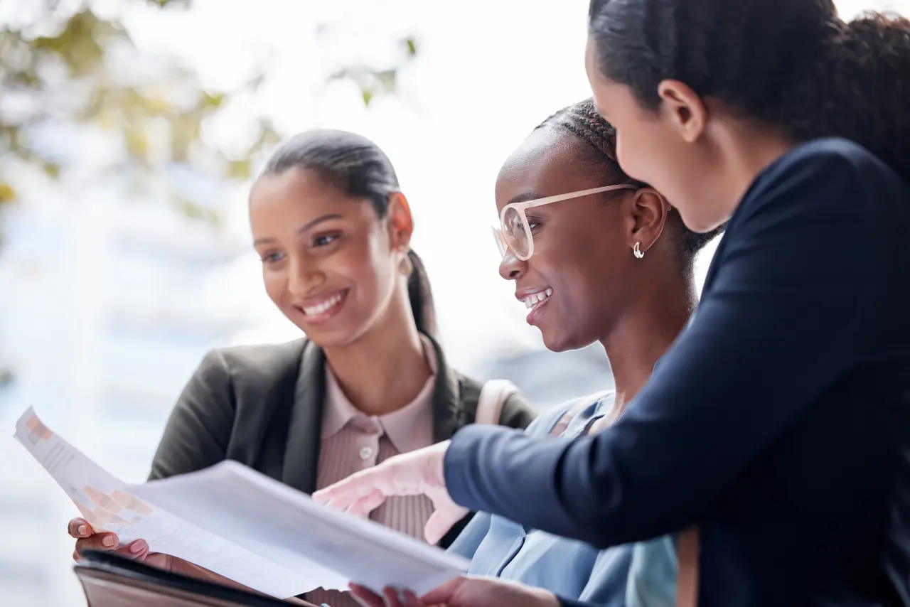 Three women engaged in discussion, reviewing documents, smiling, in an outdoor setting with greenery and urban background.