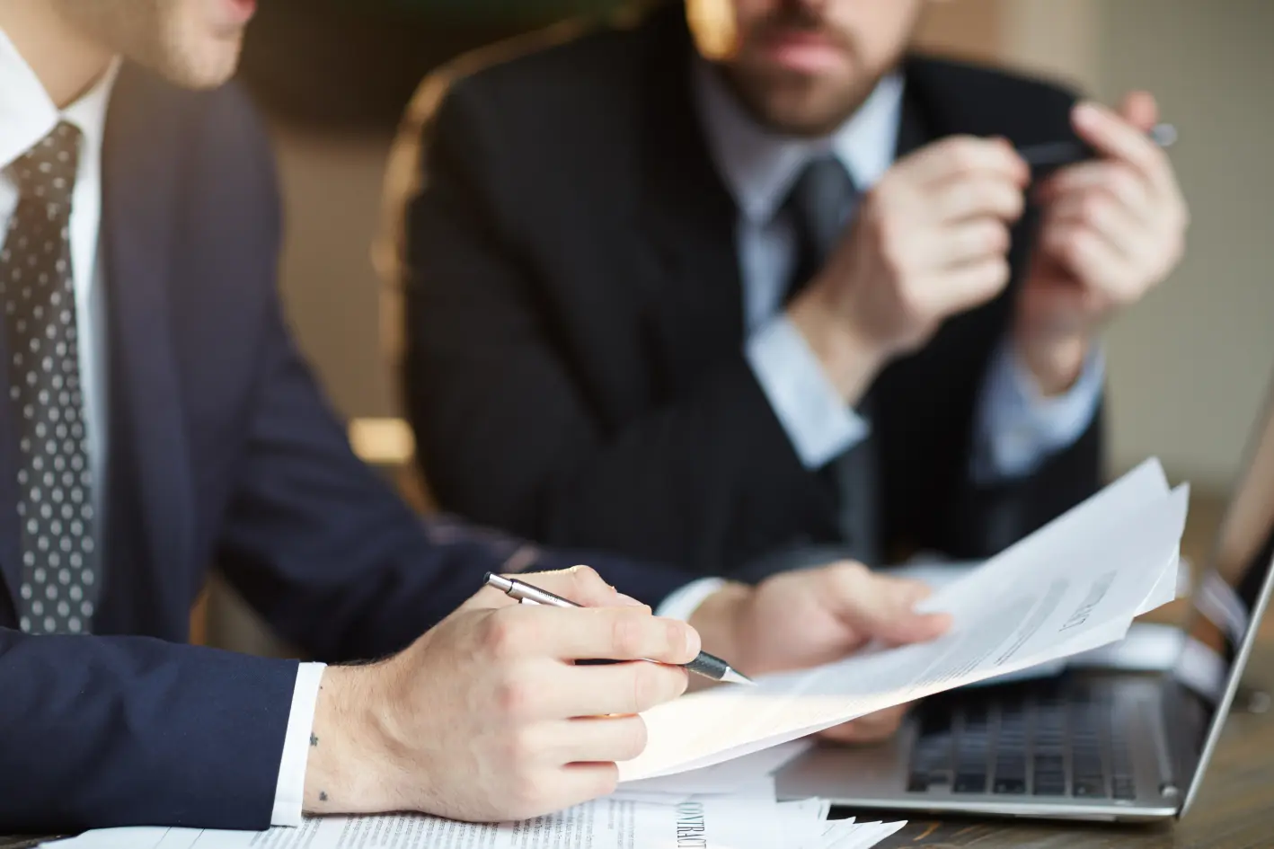 Two men in suits are discussing documents at a table, with a laptop nearby. One is taking notes with a pen.