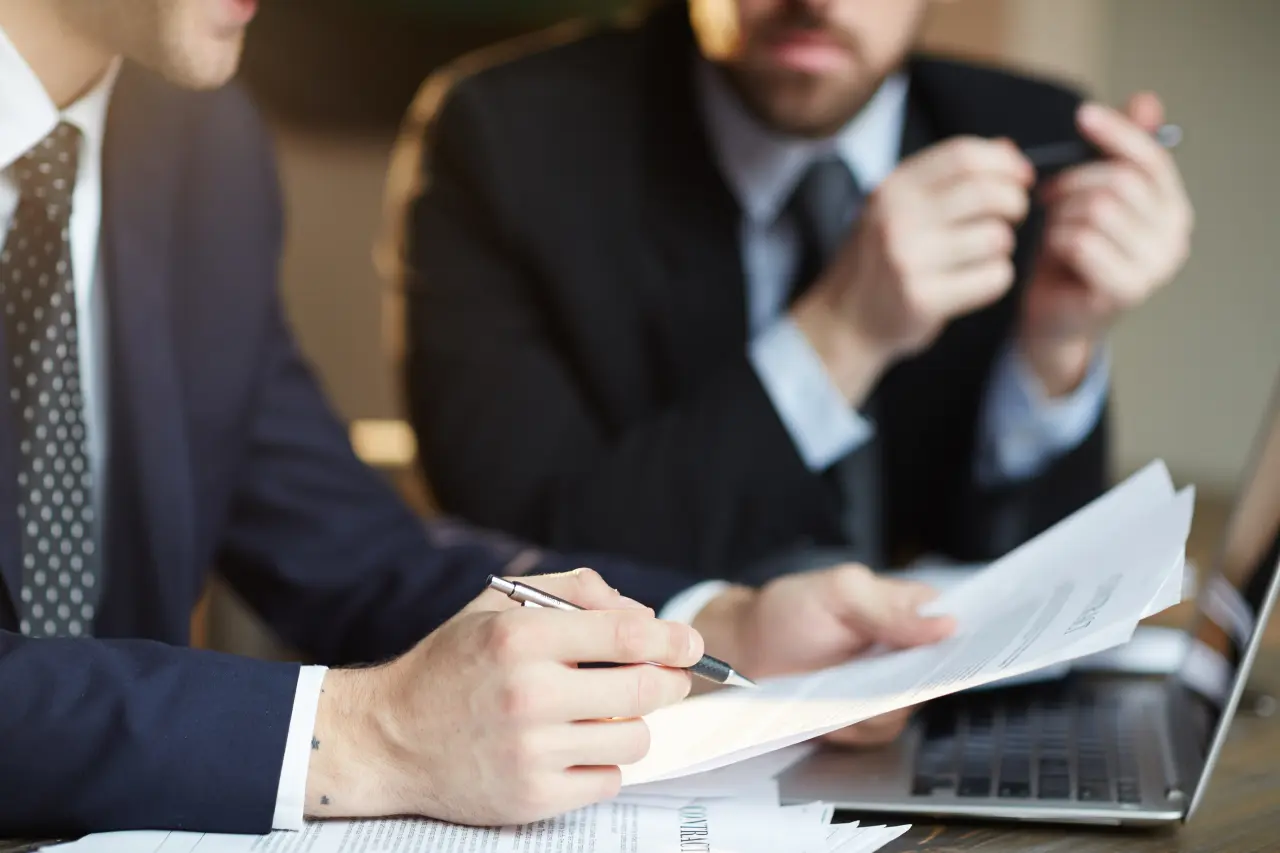 Two men in suits are discussing documents at a table, with a laptop nearby. One is taking notes with a pen.