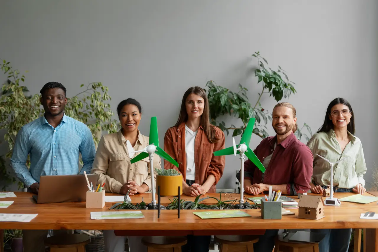 Five diverse individuals stand behind a table with models of wind turbines, plants, and a laptop, smiling in a bright indoor space.