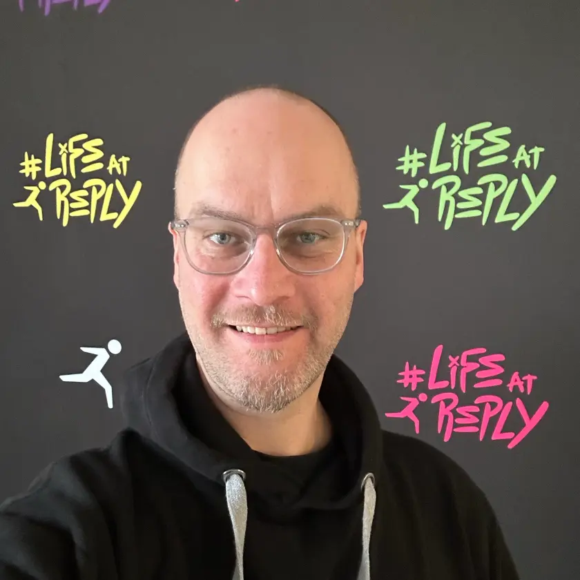 A smiling man with glasses wears a black hoodie, posing in front of a backdrop featuring colorful '#Life at Reply' graphics.