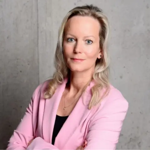 A woman with long, light blonde hair wearing a pink blazer stands confidently with her arms crossed against a concrete wall.