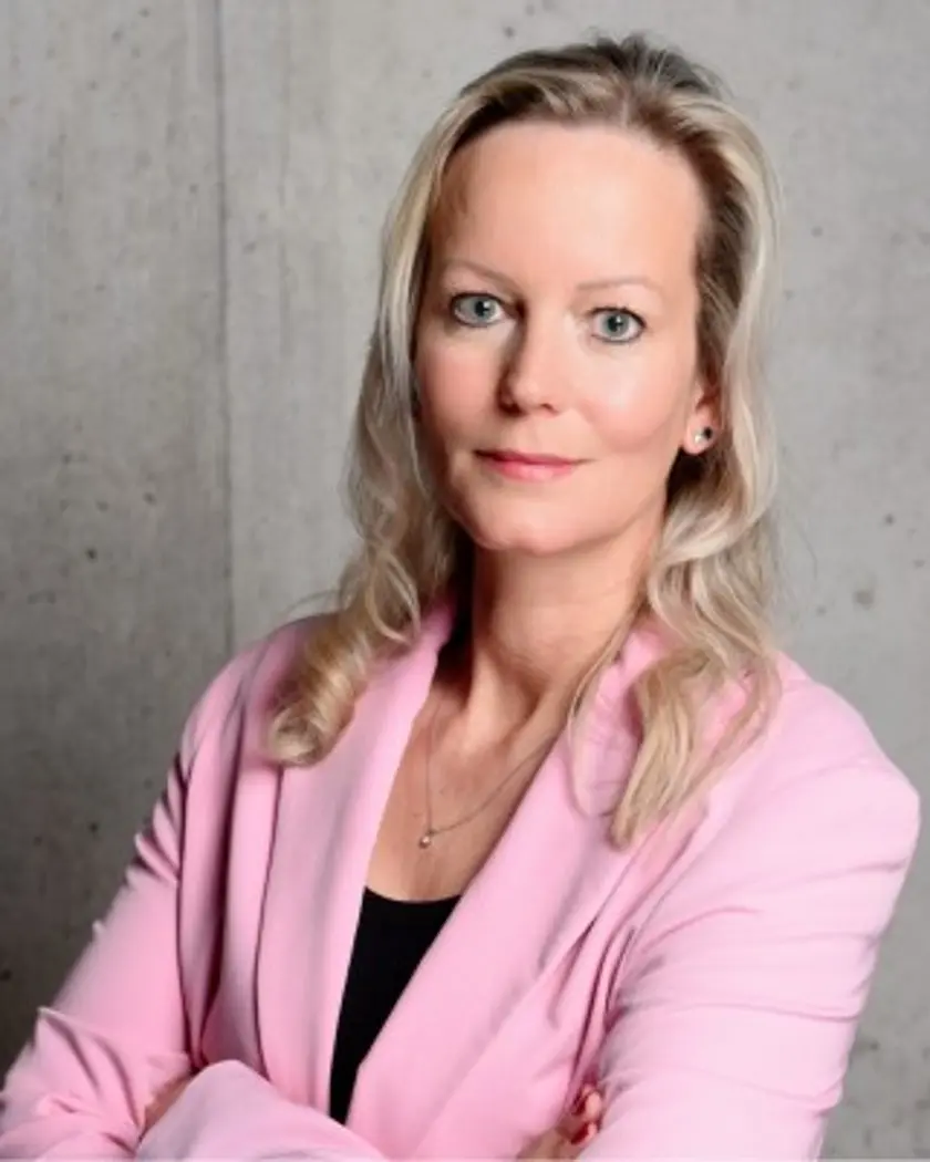 A woman with long, light blonde hair wearing a pink blazer stands confidently with her arms crossed against a concrete wall.