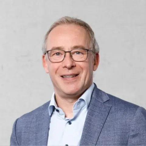 A middle-aged man with glasses and light hair smiles, wearing a blue suit jacket over a collared shirt, against a neutral background.