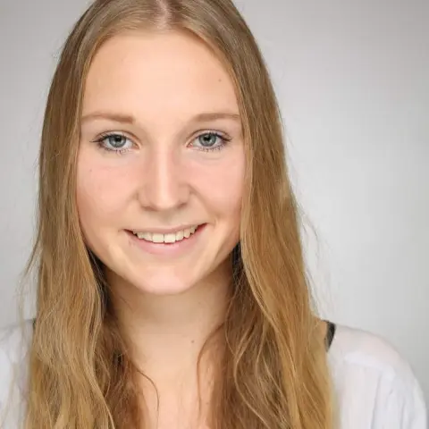 A young woman with long, straight blonde hair smiles warmly at the camera against a neutral background.