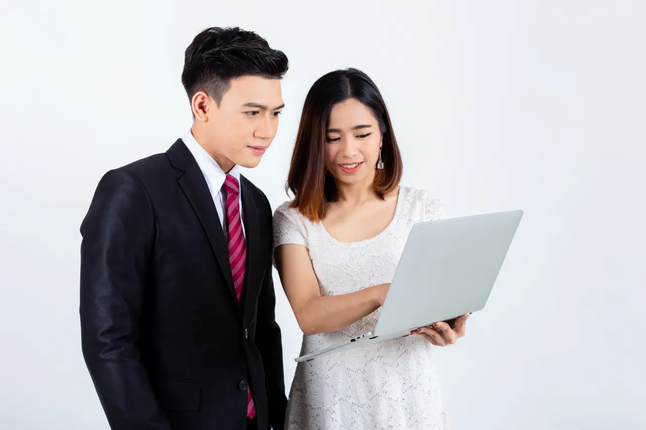 A man in a suit and a woman in a white dress are collaborating on a laptop, appearing focused and engaged.